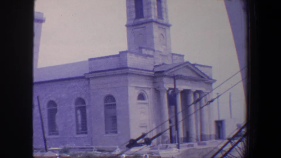 1964 Saint Louis home-movie frame showing a stone church exterior with a bell tower, arched windows, columned entrance, and scaffolding in front.