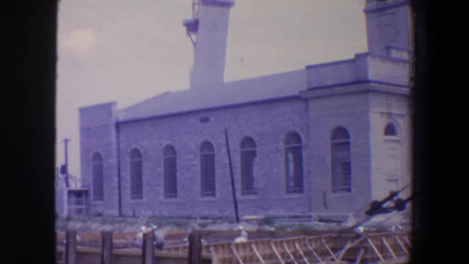 1964 Saint Louis home-movie frame showing the long side of a stone church, repeated arched windows, part of the tower, and scaffolding along the foreground.