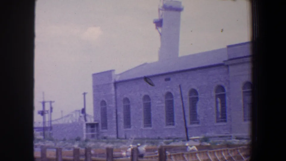 1964 Saint Louis frame showing the church side wall with arched windows, construction fencing or scaffolding, utility poles, and cars near the street.