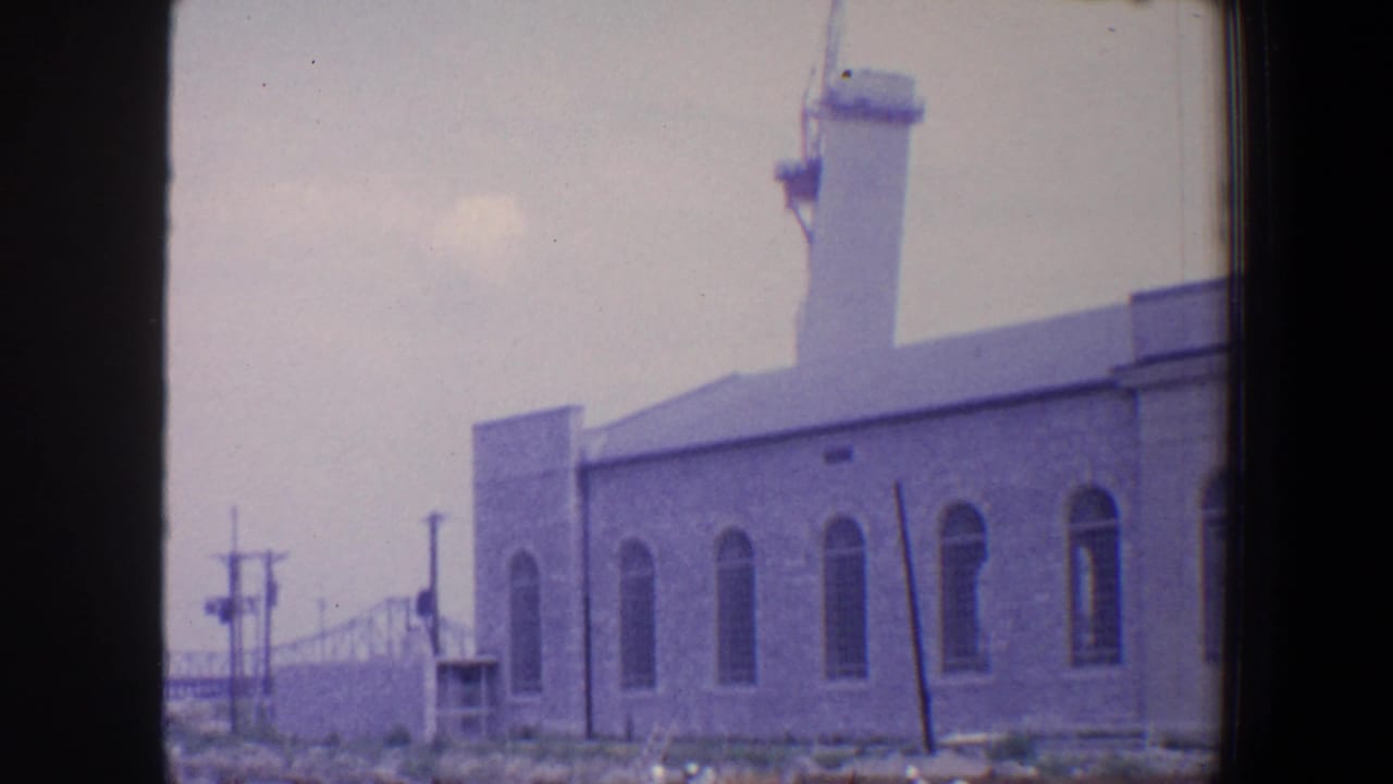 1964 Saint Louis home-movie frame showing a towered industrial building with arched windows and utility poles in front.