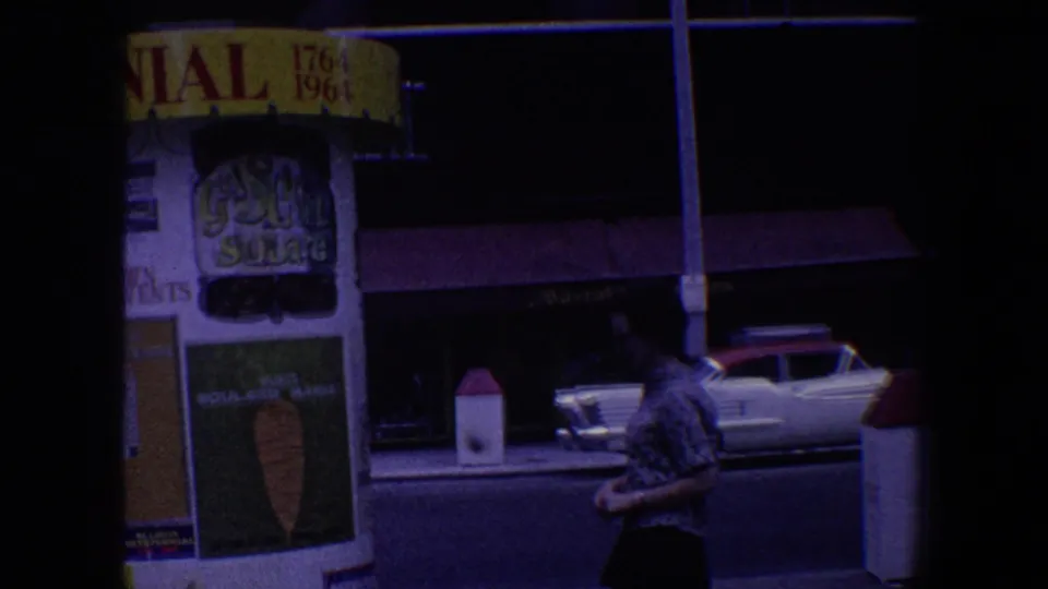 1964 Saint Louis street scene with illuminated commercial signage, storefront awnings, a classic American car, and a man walking in the foreground.