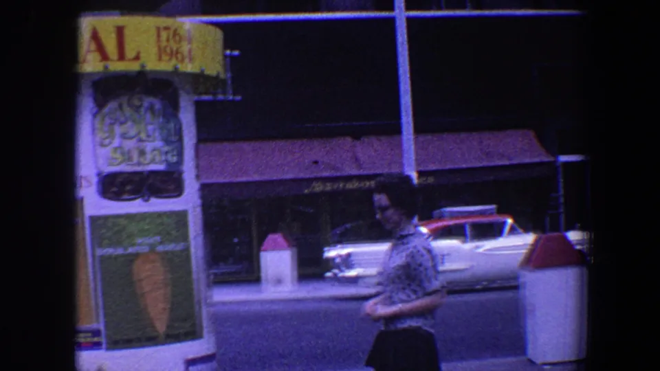 Saint Louis 1964 home-movie frame of a man walking past a classic car, street bollards, storefronts, and illuminated commercial signage.