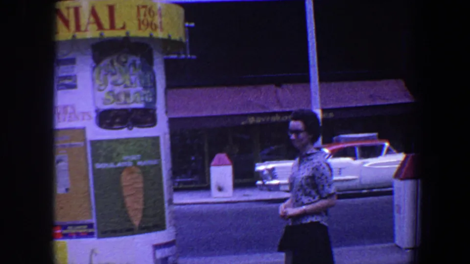 1964 Saint Louis street view with a woman passing a Centennial booth, posters, storefronts, and a parked mid-century car.