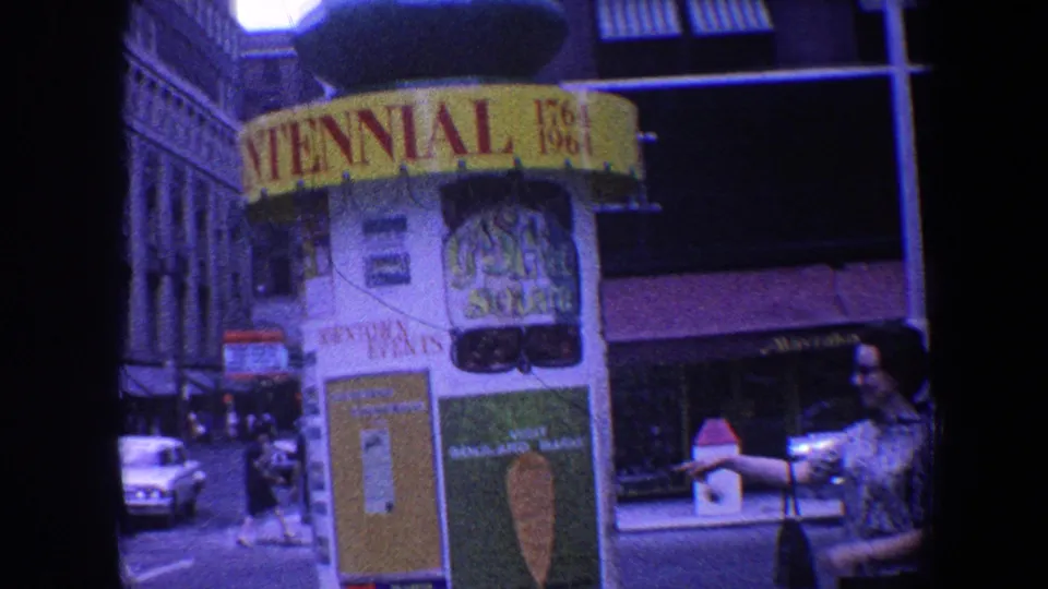 Saint Louis Centennial booth seen from the street, with civic posters, tall buildings, cars, and a woman partly visible at right.