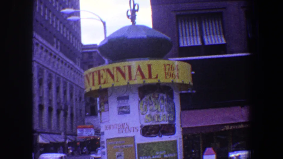 1964 Saint Louis street frame showing the Centennial booth, civic posters, a storefront awning, cars, and tall buildings in the background.