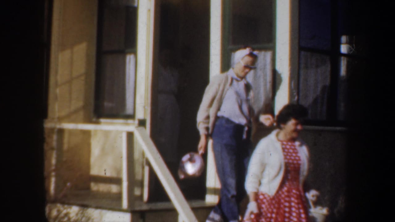 1957 Wyoming home-movie frame showing two women on porch steps, one in a red polka dot dress and another in a headscarf with a bowl.