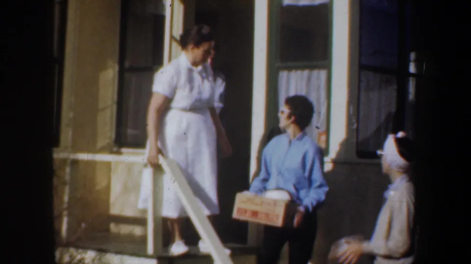 1957 Wyoming home-movie frame showing a man in glasses holding a labeled box on the steps, with two women nearby on the porch.
