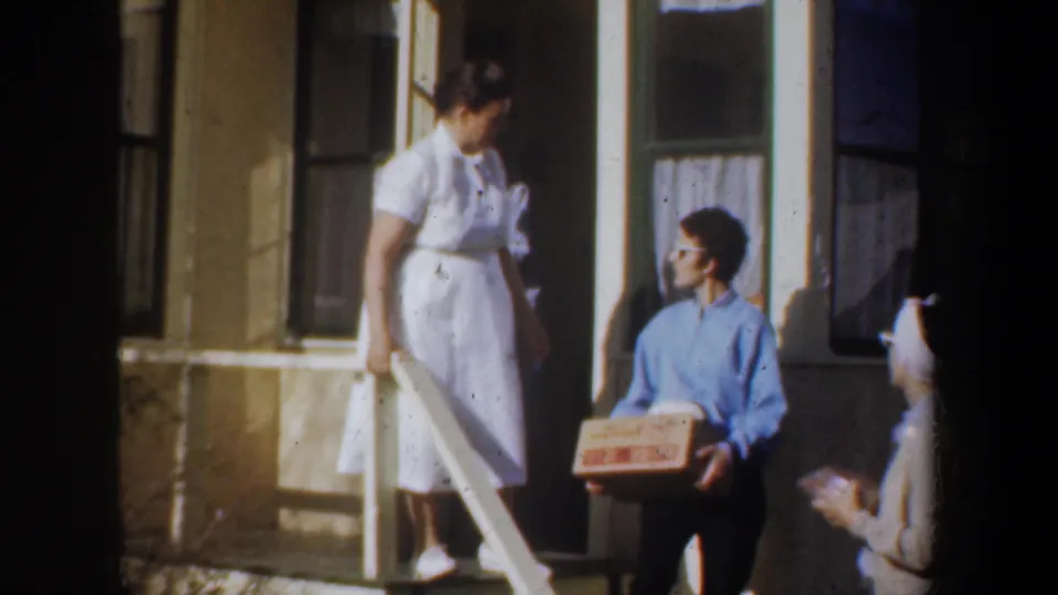 1957 Wyoming home-movie frame of a man in a blue shirt holding a labeled box while a woman in a white dress stands on the porch steps.