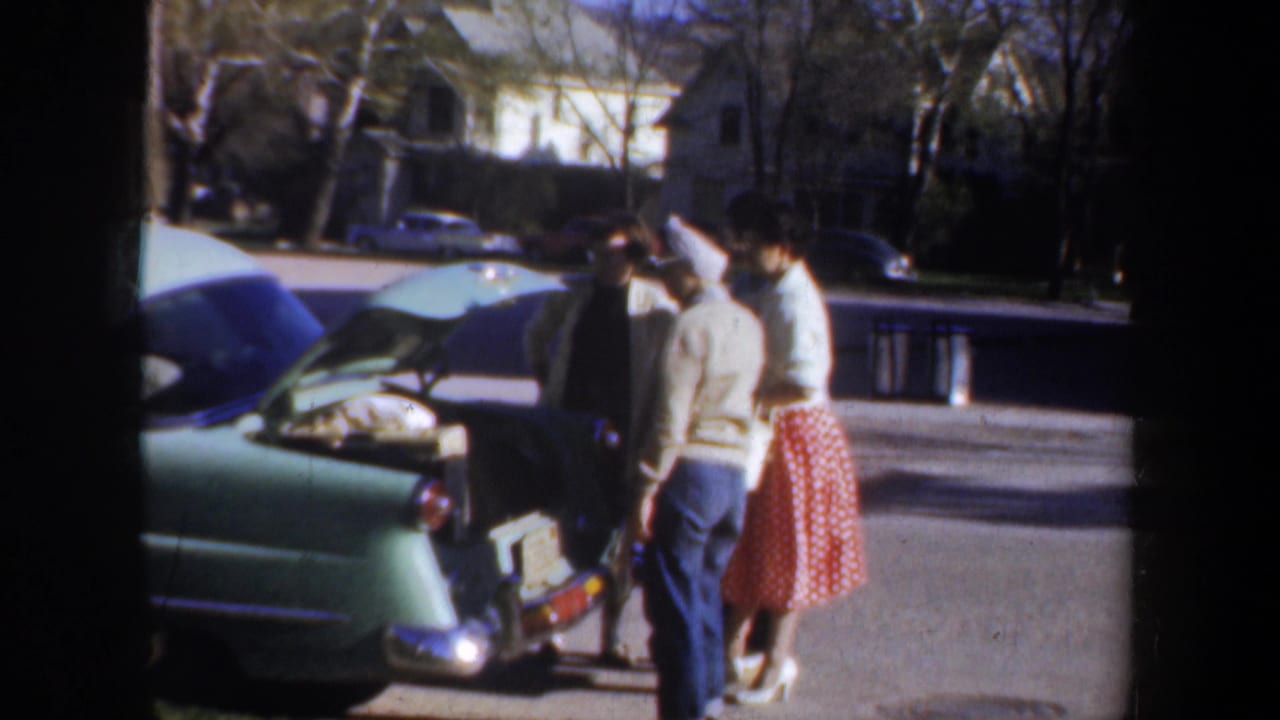 1957 Wyoming home-movie frame showing three adult women gathered by the open trunk of a light green car in a suburban neighborhood.
