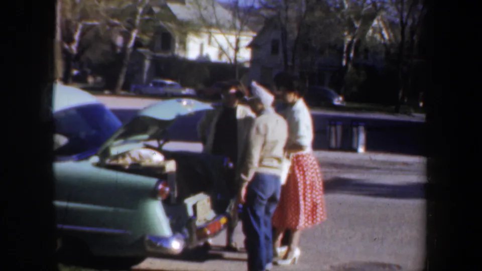 1957 Wyoming home-movie frame showing three adult women gathered by the open trunk of a light green car in a suburban neighborhood.