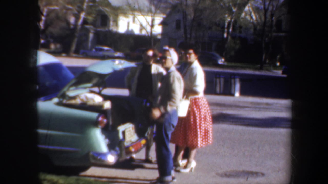 Women stand beside the rear of a light green car with its trunk open.