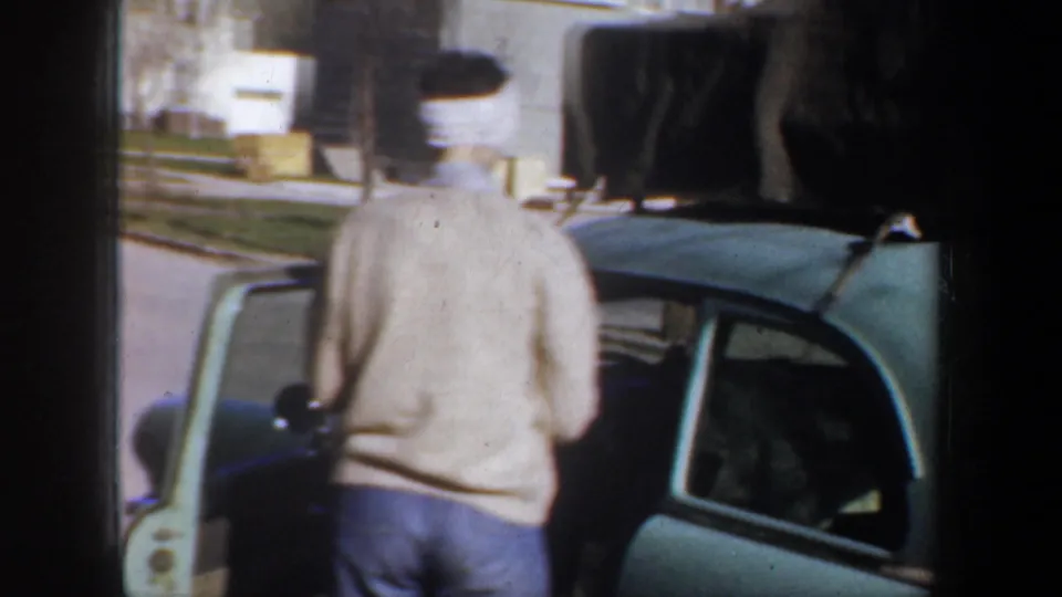 A woman in a light sweater stands near an open door of the green car, with bundled luggage visible on the roof.
