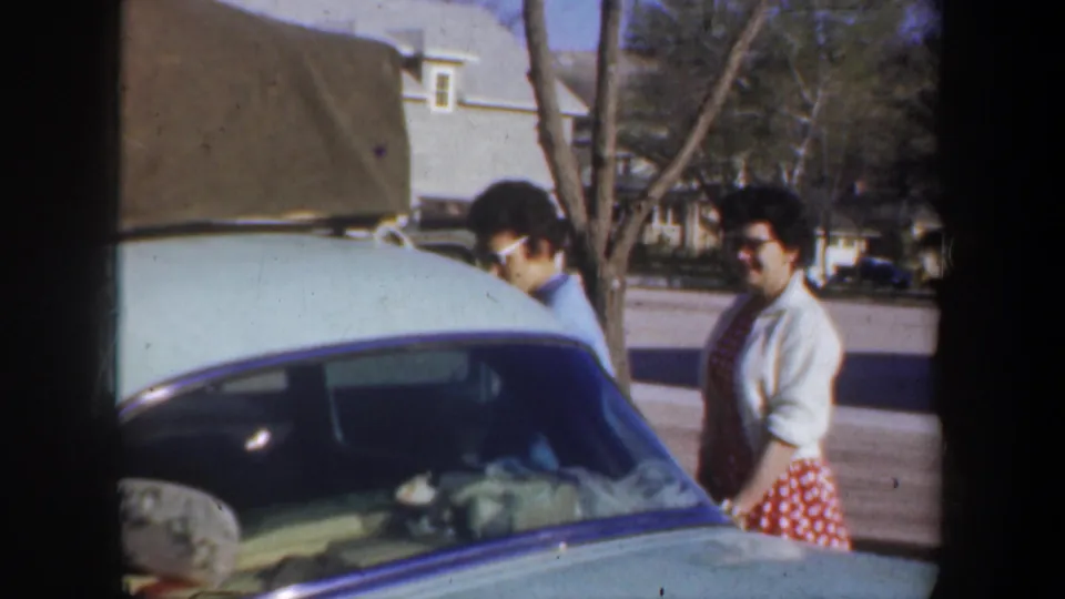 Two adult women stand near the side of the light green car, one in a red polka dot skirt, with roof luggage above them.