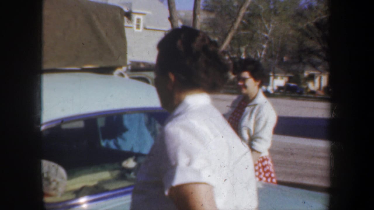 Wyoming 1957 home-movie frame showing adults near a pale car in a residential street as road-trip preparations appear underway.