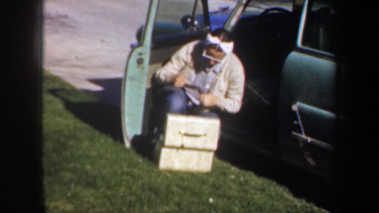 Woman kneels by an open car door with a small suitcase on the grass.