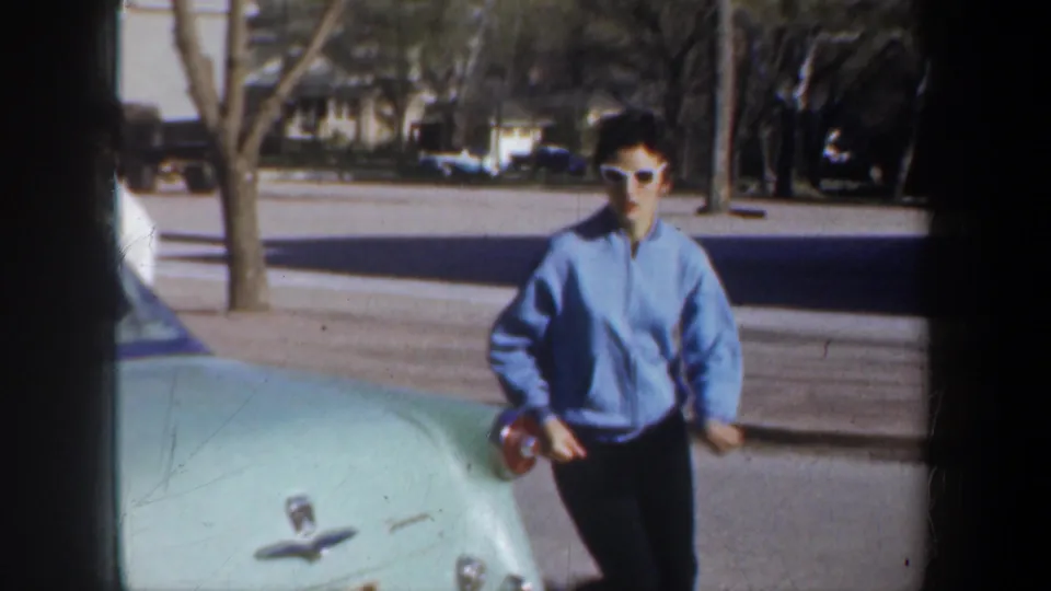 Wyoming 1957 home-movie frame showing a person in a blue jacket near the hood of a pale car with houses and trees beyond.