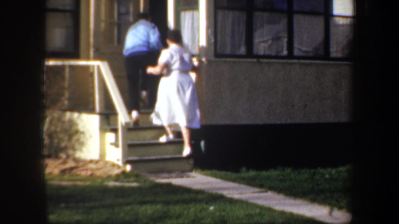 In 1957 Wyoming home-movie footage, a man in a blue jacket and a woman in a white dress climb steps to a house entrance.