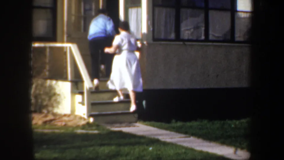 In 1957 Wyoming home-movie footage, a man in a blue jacket and a woman in a white dress climb steps to a house entrance.
