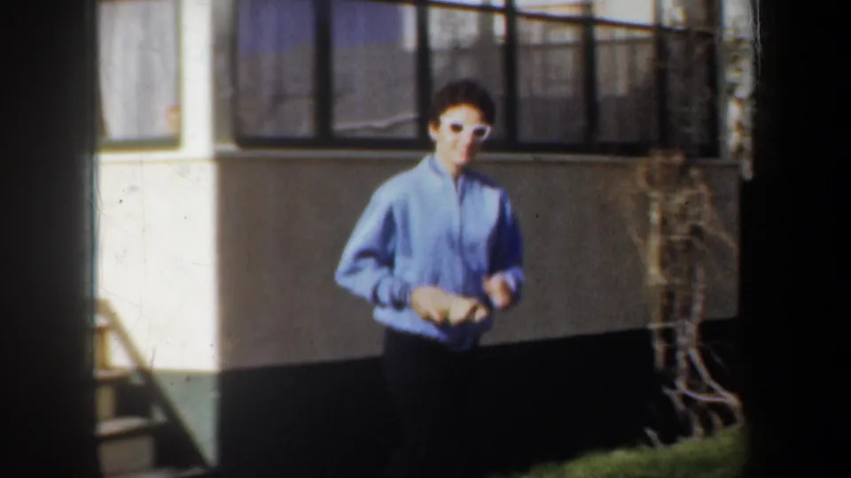A man in a blue jacket stands near the exterior wall and curtained windows of a Wyoming house in 1957 footage.