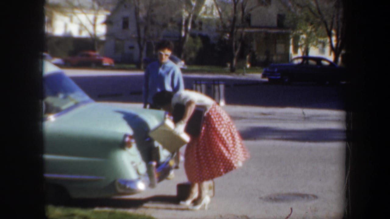 Woman bends near the rear of a green car while a man stands behind her on a residential street.