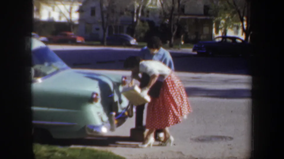 A woman in a red skirt holds a box at the back of the green car while the man in blue leans closer to assist.