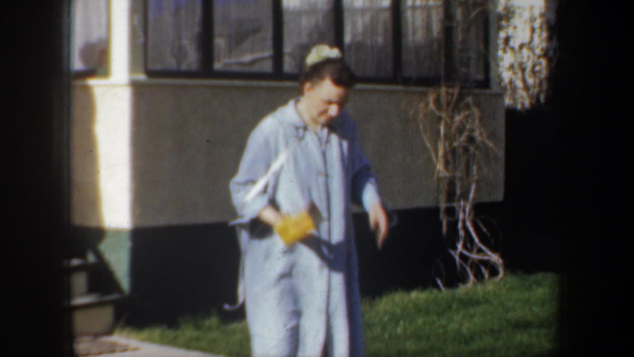 In 1957 Wyoming home-movie footage, a woman in a light blue dress walks by a house while holding a bright yellow box.