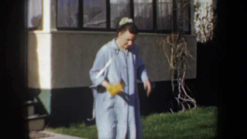 In 1957 Wyoming home-movie footage, a woman in a light blue dress walks by a house while holding a bright yellow box.