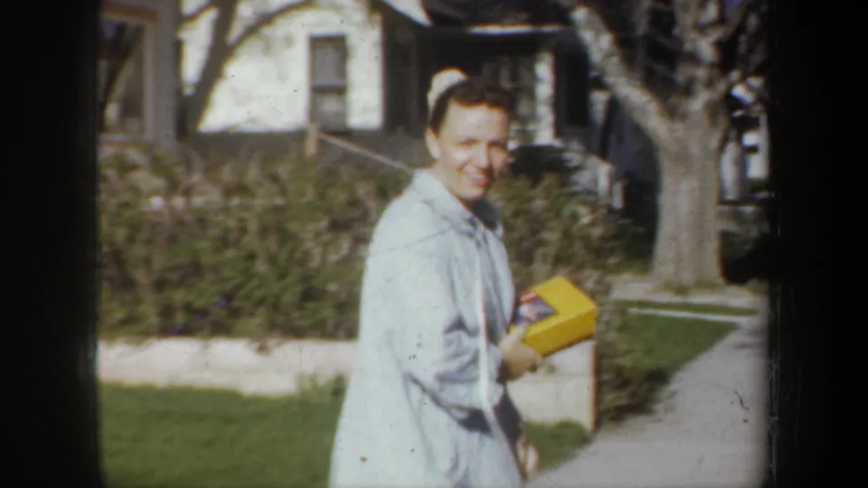 A woman in a light blue dress smiles toward the camera while holding a yellow box on a residential sidewalk in Wyoming, 1957.