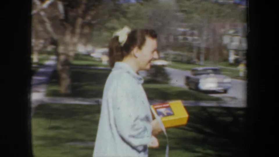 Side view of the woman carrying a yellow box near lawns, trees, and parked cars on a Wyoming residential street in 1957.