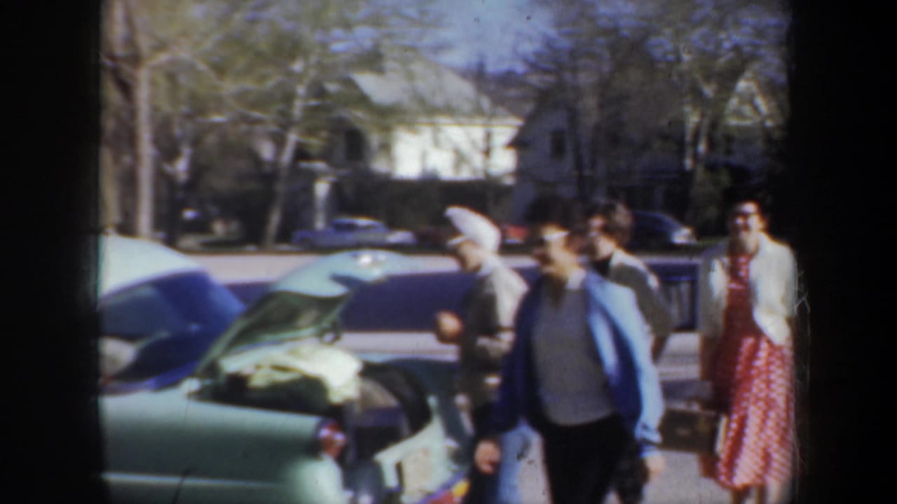 Adults gather beside a light green car with its trunk open on a residential street.