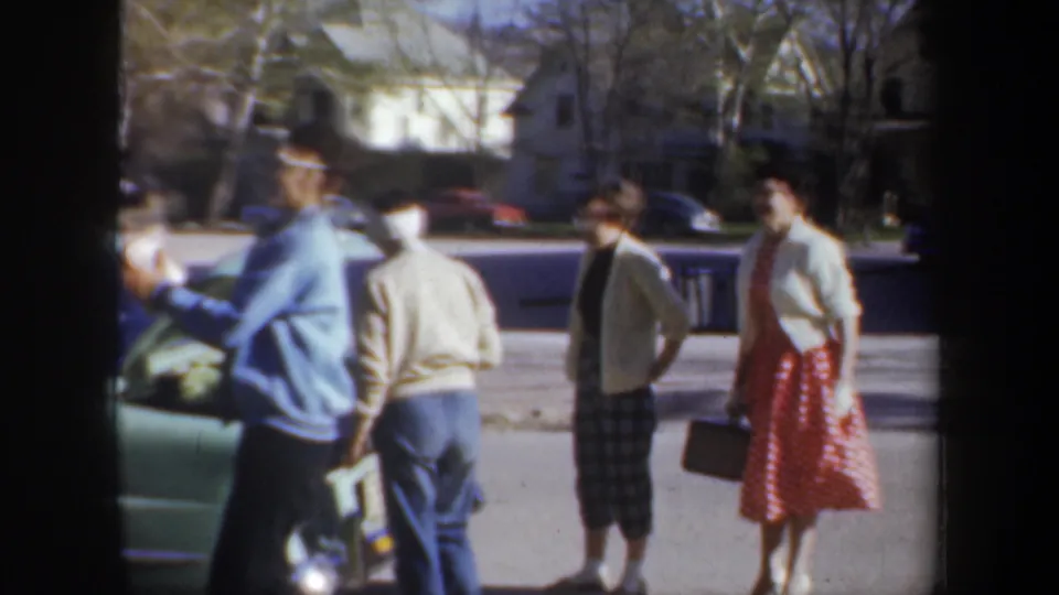 A small group stands by an open car trunk, including a woman in a red polka-dot dress and others in casual 1950s clothing.