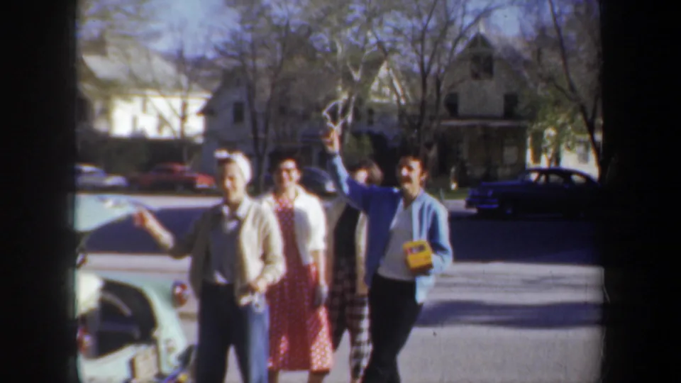 Several adults smile and wave beside a car in a 1957 Wyoming suburban home-movie scene.
