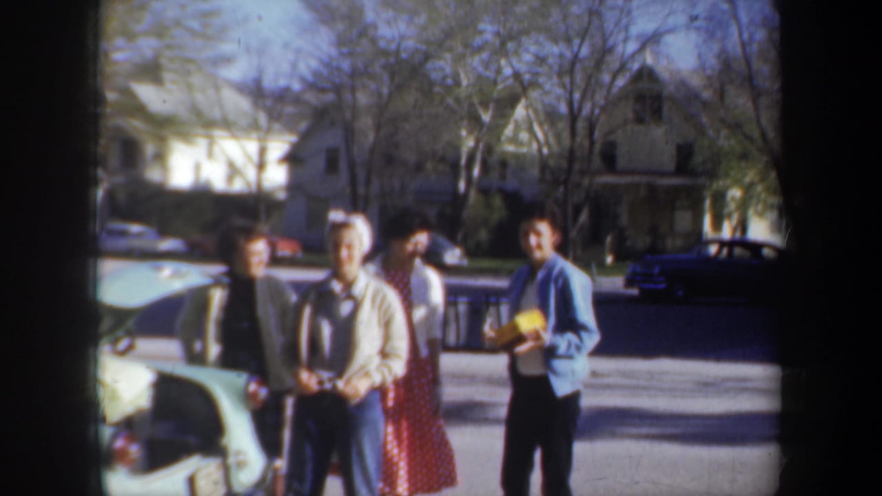 Four young adults stand beside a pale car on a sunny residential street.
