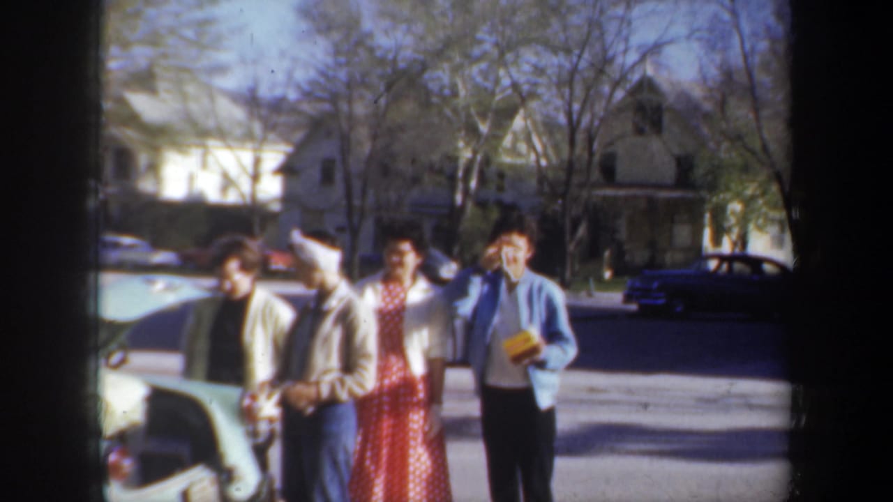 A young man in a blue sweater waves while holding a yellow box near three others.