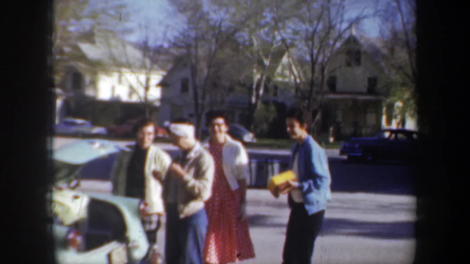 1957 Wyoming home-movie frame showing four young adults conversing near a car, with houses and parked vehicles in the background.