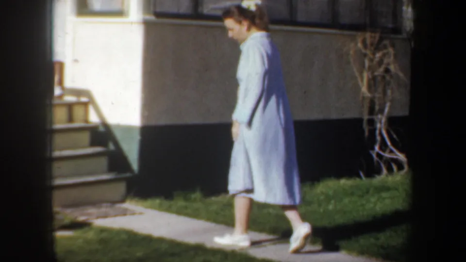 1957 Wyoming home-movie frame of a woman in a light blue dress walking past porch steps and a lawn.