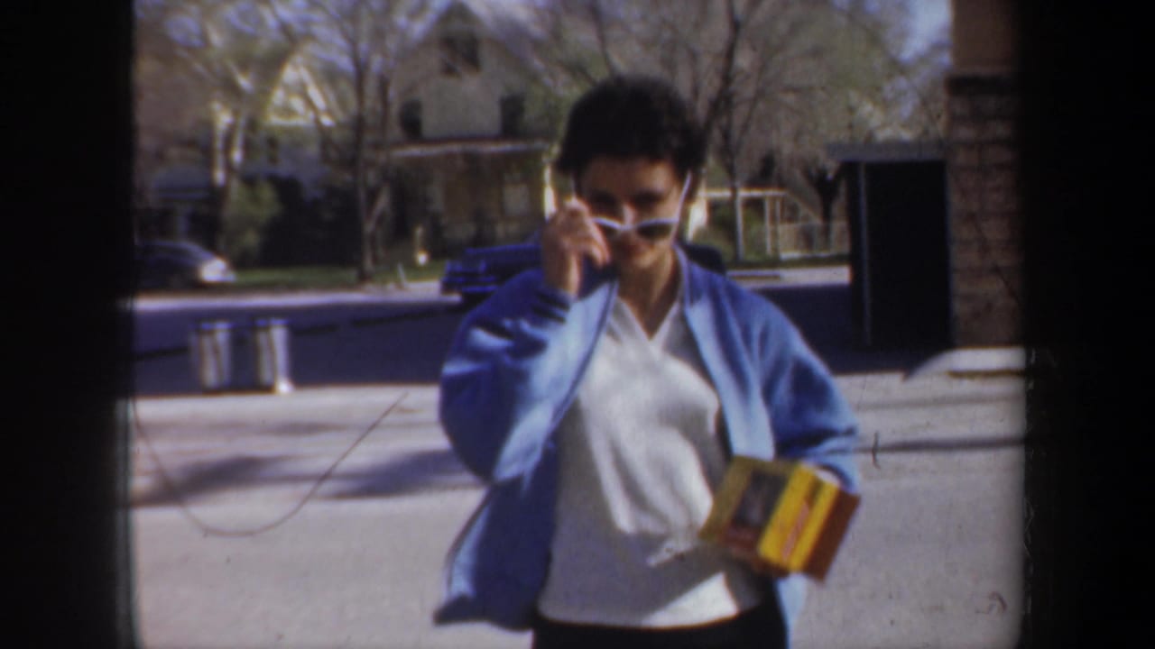 Woman in blue jacket adjusts sunglasses while walking toward the camera outdoors.
