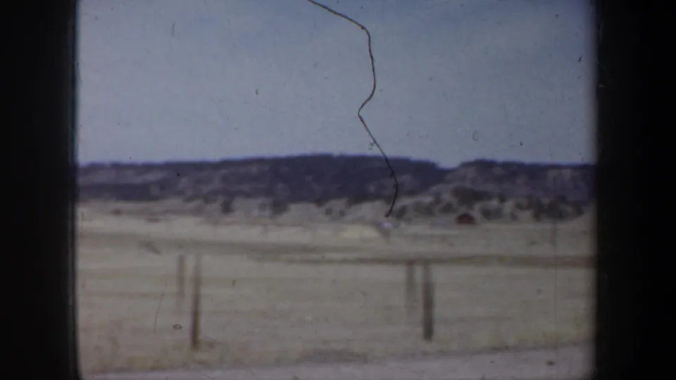1957 Wyoming roadside view from a moving vehicle, showing dry ground, fence posts, low hills, and pale sky.