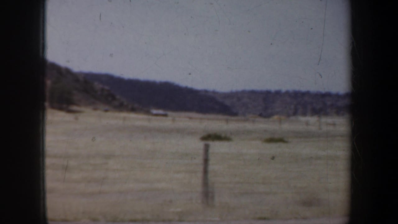 Open field with a fence post, low hills, and cloudy sky seen through dark car-window framing.