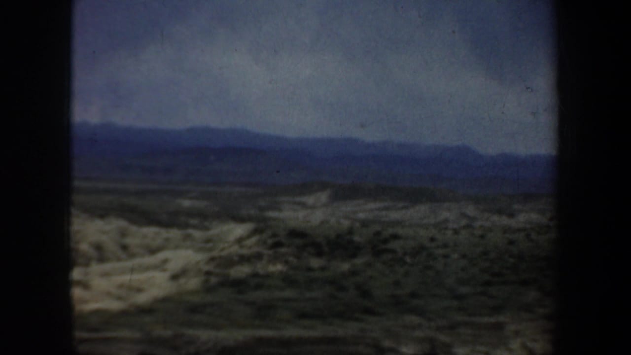 Scrubby rolling landscape and distant mountains under a gray sky, framed by dark film or window edges.