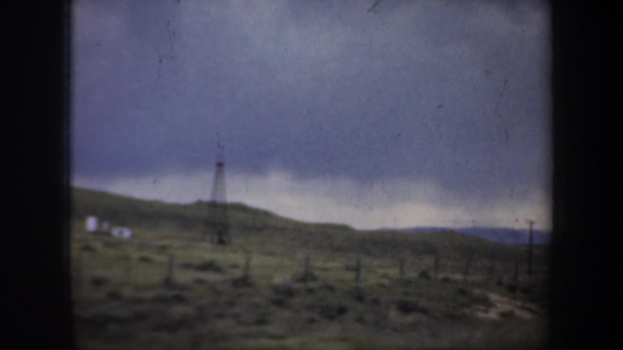 Oil derrick on a low hill with utility poles and open ground under a dark sky.