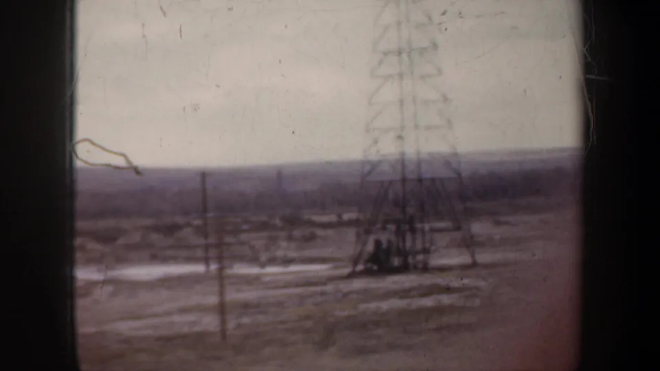 1957 Wyoming 8mm frame showing a transmission tower, utility lines, and oil-field equipment across a sparse plain.