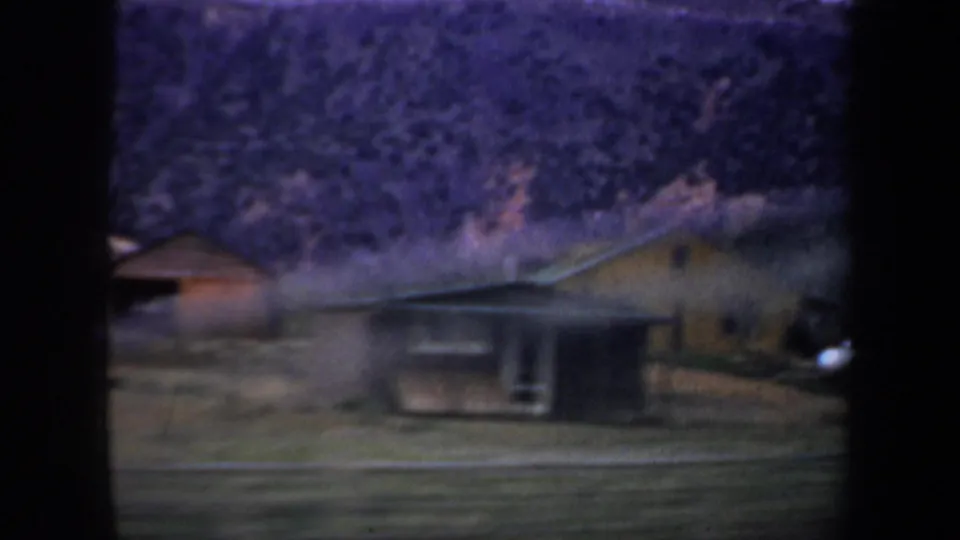 1957 Wyoming home-movie frame with rural buildings near a road, backed by a steep scrub-covered hillside.