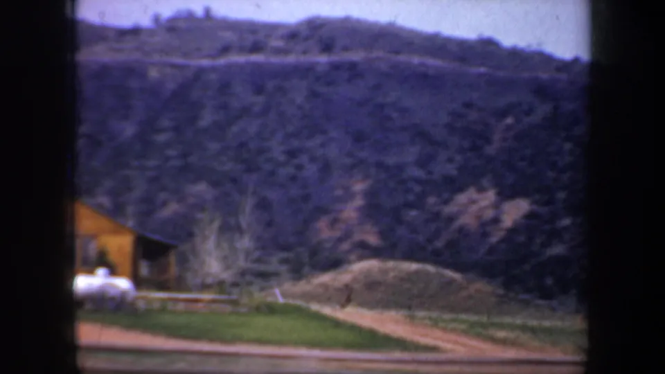 Wyoming 1957 8mm frame showing a small rural building beside a dirt road with a mountain slope in the background.