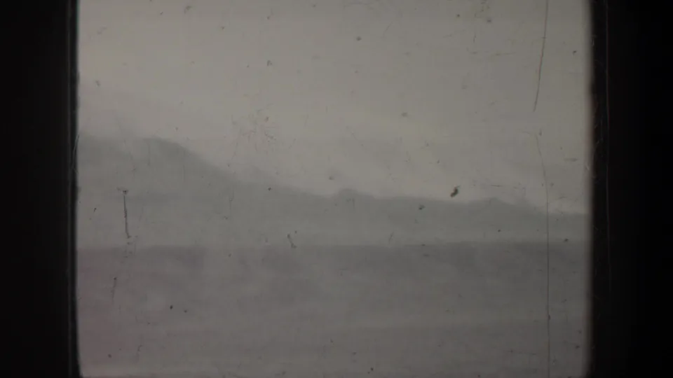 1957 Wyoming 8mm test frame with a hazy mountain landscape, pale sky, dark borders, dust, and a faint pole-like mark.