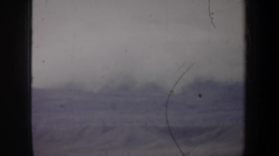 1957 Wyoming 8mm frame showing distant low terrain beneath a pale sky, with heavy film wear and dark gate edges.