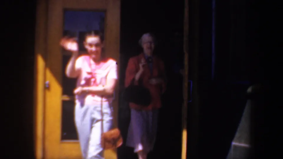 Two adults stand at yellow doorways in bright and shadowed light, one waving toward the camera.
