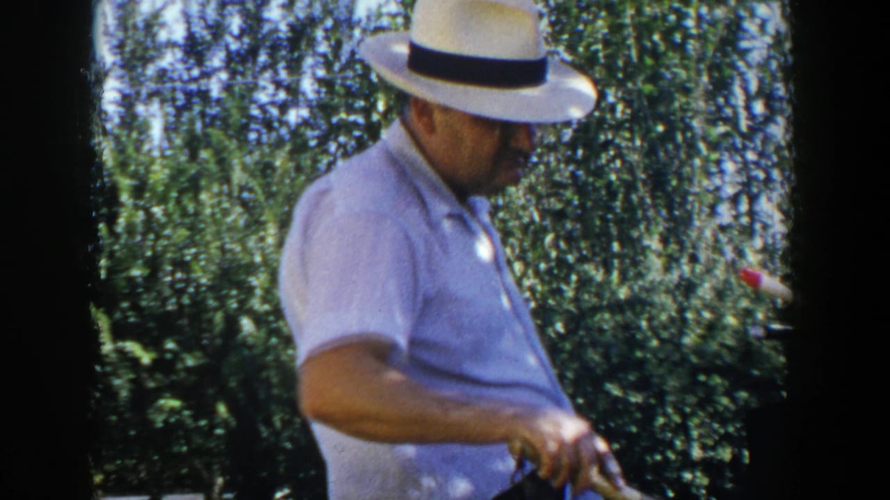 Man in a white fedora stands by an outdoor grill in a sunny backyard.