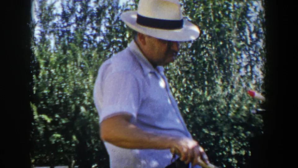 In 1953 Lubbock, Texas home-movie footage, a man in a white fedora tends a backyard grill beside shrubs and lawn.