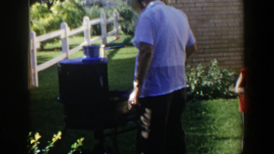 A man stands near an outdoor grill with a pot on top in a Lubbock backyard, with a white fence and brick wall visible.